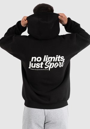 Persoon die een zwarte hoodie met de tekst "no limits, just Sport" op de rug draagt, de capuchon verstelt, grijze broek, tegen een effen lichte achtergrond.
