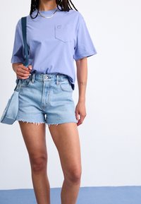 Maglietta a maniche corte color lilla chiaro con taschino sul petto, abbinata a pantaloncini di jeans azzurri con bordi sfilacciati e design a cinque tasche.