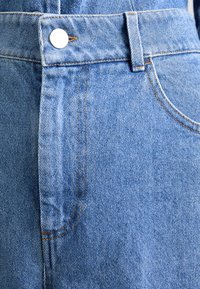 Ljust blå denimtyg med synlig sömnad, en silverknappstängning och en högmidjad skärning med fram- och sidofickor.