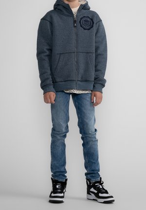 Grey zip-up hoodie with a textured pattern and navy emblem, paired with light blue jeans and black and white sneakers.