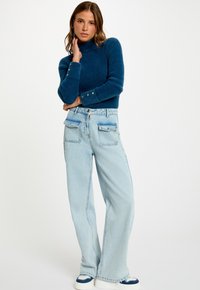 Maglione blu a collo alto con maniche lunghe e dettagli con bottoni, abbinato a jeans a gamba larga blu chiaro con tasche a patta.