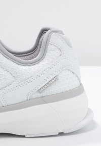 White sneaker featuring mesh upper with circular pattern, grey accents, padded collar, and a rubber sole. Logo displayed on the side.