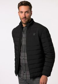 Giacca nera imbottita con colletto alto, caratterizzata da un logo sottile sul davanti. Indossata sopra una camicia a quadri grigia e abbinata a jeans grigi.