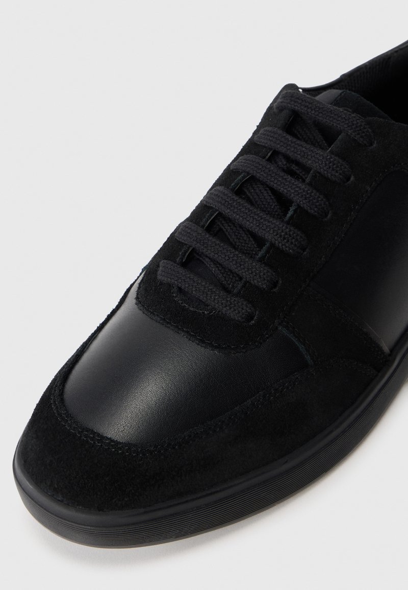 Zapatilla negra que presenta una combinación de cuero liso y gamuza texturizada. Diseño de cordones con puntera redondeada y suela de goma para mayor durabilidad.