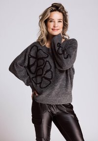 Grauer übergroßer Pullover mit schwarzer Blumenstickerei. Gerippte Bündchen und Saum, kombiniert mit glänzend schwarzen Hosen mit seitlichen Taschen.