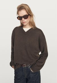 Hnědý svetr s výstřihem do V a měkkou texturou, doplněný tmavými džínami a mohutnými hnědými slunečními brýlemi. Model stojí uvolněně s rukama v kapsách.