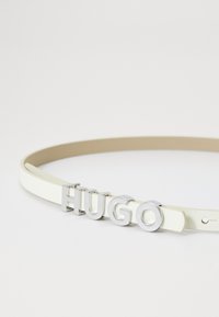 Weißer Leder-Gürtel mit einem silbernen Metall-Logo, das vorne "HUGO" spellt. Der Gürtel hat eine glatte Textur und ein minimalistisches Design.