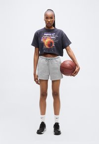 Αποκομμένη μαύρη γραφική μπλούζα με σχέδιο μπάσκετ; ανοιχτό γκρι σορτς με κορδόνι; μαύρα αθλητικά παπούτσια με λευκές λεπτομέρειες; κρατώντας μια μπάλα μπάσκετ.