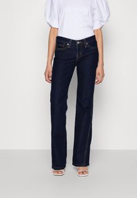 Jean bleu foncé avec une coupe droite, cinq poches et des détails de couture jaunes. Associé à un haut à manches bouffantes blanc et des sandales blanches.