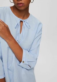Femme portant un chemisier bleu clair à manches longues avec manches retroussées et col à nouer, accessoirisé avec des boucles d'oreilles argentées en forme d'anneaux.