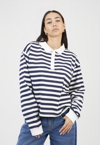 Woman with dark hair wearing gold hoop earrings, a navy and white striped long-sleeve polo shirt, and blue jeans, with one hand in pocket.