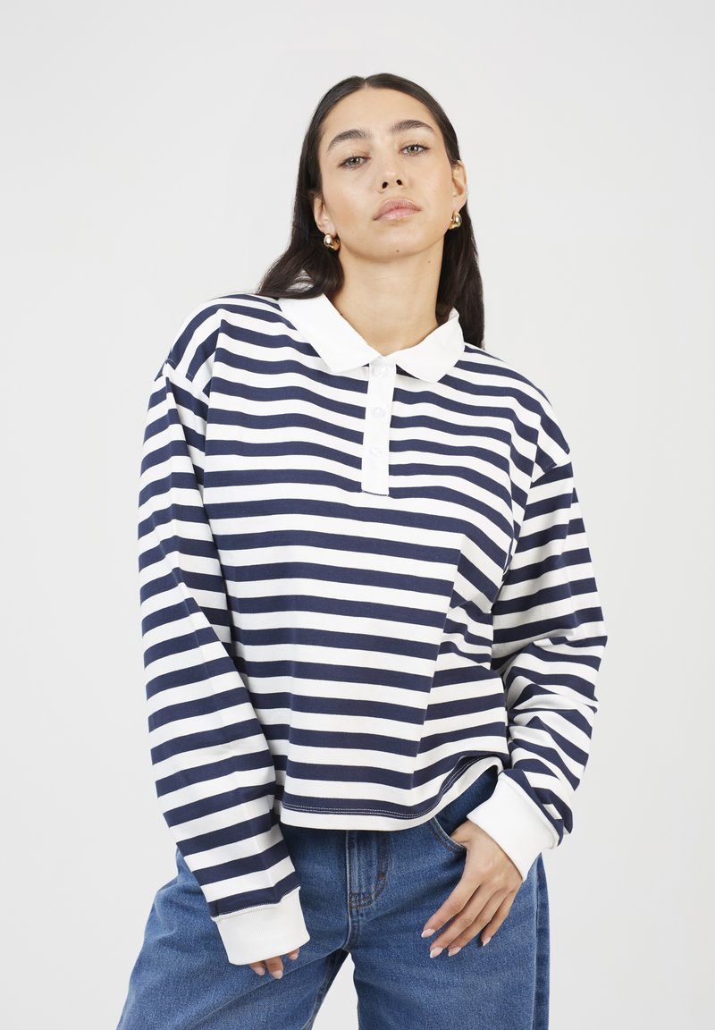 Woman with dark hair wearing gold hoop earrings, a navy and white striped long-sleeve polo shirt, and blue jeans, with one hand in pocket.