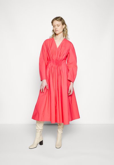 Vestido de algodão coral com decote em V profundo, cintura franzida, mangas longas e saia godê. Combinado com botas de cano alto em creme e um fundo minimalista.