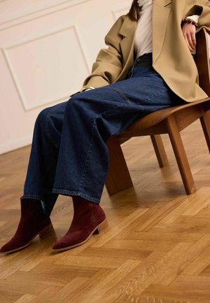 Person trägt einen beigen Blazer, ein weißes Oberteil, weit geschnittene blaue Jeans und bordeauxrote Wildleder-Stiefeletten und sitzt auf einem Holzstuhl auf Parkettboden.