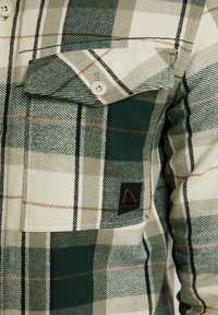 Chemise en flanelle à carreaux verts et crème avec étiquette noire, comprenant une poche à boutons et un détail de couture contrastant orange.