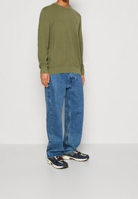 Mężczyzna w oliwkowo-zielonym swetrze z długim rękawem, luźnych niebieskich dżinsach i wielokolorowych tenisówkach, stojący na jasnoszarej podłodze i tle.