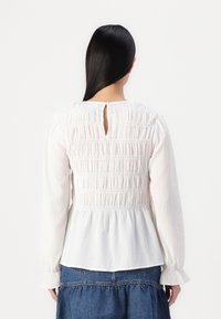 Hvit langermet bluse med teksturert smocking på baksiden og en nøkkelhulls åpning. Mykt stoff med en bølgende nederkant og rynkede mansjetter.