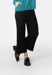 Schwarze, gerippte, weit geschnittene Hosen mit gewelltem Saum, getragen mit schwarzen Plateaustiefeln. Das Oberteil hat eine einheitliche petrolfarbene Farbe und einen lässigen Schnitt.