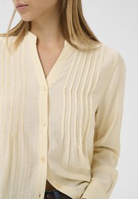 Lichtgele, knoopsluiting blouse met subtiele verticale plooien en een mandarin kraag. Zachte, gestructureerde stof met lange mouwen.