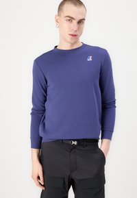 Paarse longsleeve sweater met een ronde hals, voorzien van een klein logopatch op de linkerborst, gecombineerd met zwarte cargobroeken.