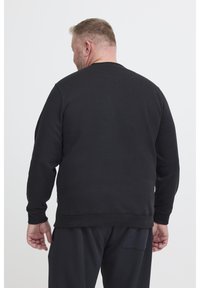 Uomo con capelli corti e barba, che indossa una felpa nera a maniche lunghe e pantaloni neri, girato di spalle su uno sfondo bianco.
