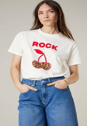 Femme portant un t-shirt blanc avec le texte rouge "ROCK" et des cerises à imprimé léopard, assorti à un jean bleu taille haute, mains dans les poches.