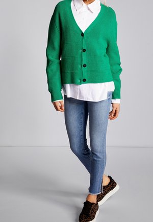 Femme portant un cardigan vert boutonné sur une chemise blanche, un jean skinny bleu et des baskets noires à motifs, debout sur un sol gris.