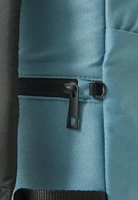Mėlyna audinio kuprinė su juodu užtrauktuku ir traukimo juostele, turinti tekstūruotą paviršių ir kontrastingą pilką medžiagą šalia.