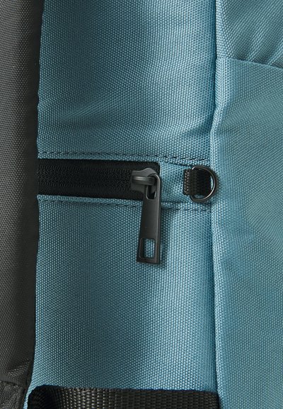 Section d'un sac à dos en tissu teal avec une fermeture éclair noire et un tirette, présentant une finition texturée et un matériau gris contrastant à côté.