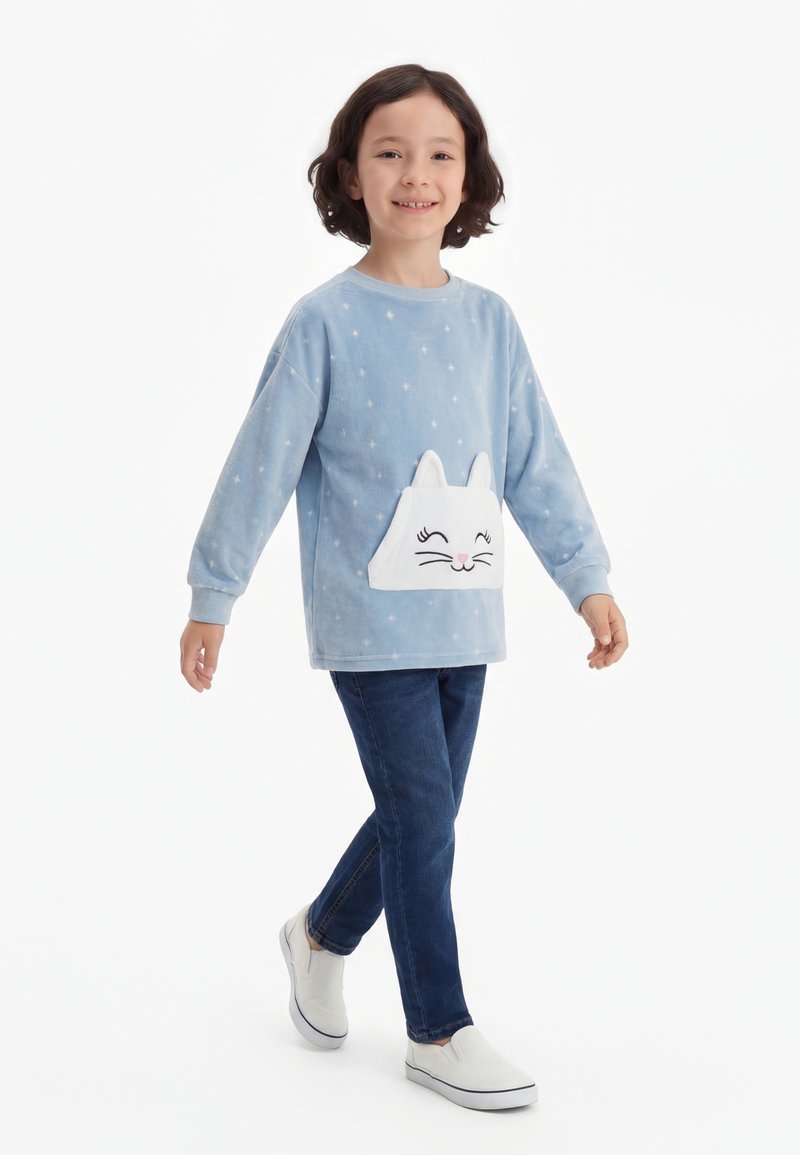 Barn går, iført en lyseblå sweater med hvidt katteansigt, mørke jeans og hvide slip-on sko, mod en ensfarvet hvid baggrund.