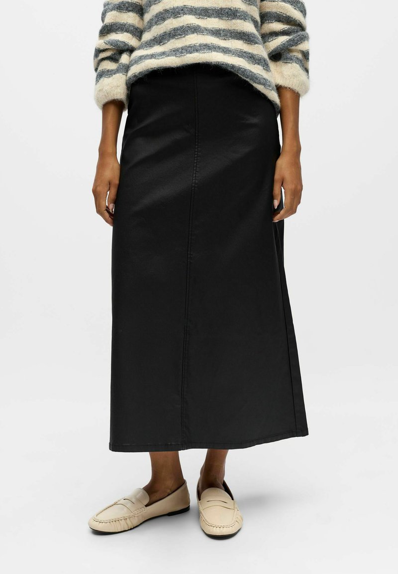 Schwarzer Baumwoll-Maxikleid mit geradem Schnitt und sichtbaren Nähten. Kombiniert mit hellbeigen Slippern. Das Modell trägt einen gestreiften Pullover.