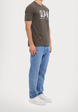 Man wearing black Armani Exchange t-shirt, light blue jeans, and white sneakers standing against plain light background.