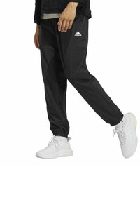 Schwarze sportliche Jogginghosen mit elastischen Bündchen und einem Adidas-Logo auf der linken Seite, kombiniert mit weißen Sneakers. Leichtes, glattes Material.