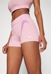 Rosa idrettsshorts med ribbestrikket liv, med et lila stripemønster og en hvit logo på benet. Glatt, elastisk stoff.