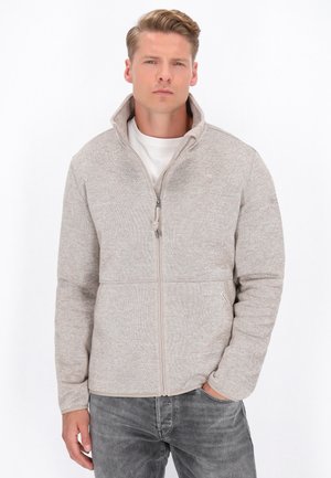 Beige Zip-Jacke aus Fleece mit hohem Kragen, strukturiertem Strickstoff und vorderen Taschen, kombiniert mit einem weißen Hemd und grauen Jeans.
