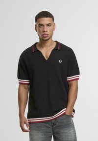 Polo negro de manga corta con un tejido texturizado, cuello en V, acentos a rayas rojas y blancas en las mangas y el dobladillo, y un pequeño logo en el pecho.