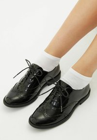 Černé lakované oxfordky s detaily ve stylu wingtip, s šněrováním a nošené s bílými kotníkovými ponožkami. Hladký a lesklý povrch.