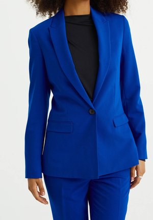 Vrouw draagt een nauwsluitend felblauw colbert met bijpassende broek en een zwart topje, staand met ontspannen handen langs het lichaam.