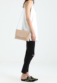 Nahá kabelka ve stylu obálky s kovovým akcentem, nošená přes tělo se stříbrným řetízkovým popruhem. Modelka ji kombinuje s černými potrhanými džíny a vyšívanými botami.