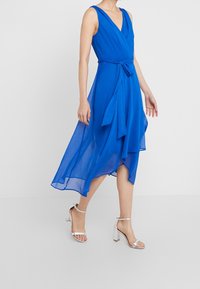 Robe portefeuille bleu sans manches en tissu léger avec un col en V et une ceinture ajustable à la taille. Elle présente un ourlet asymétrique fluide.