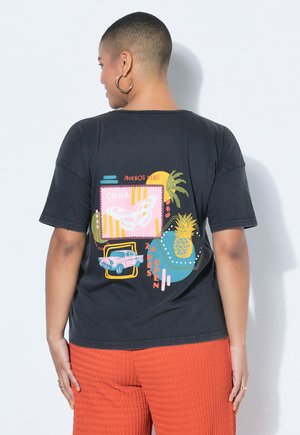 Person with short hair wearing black t-shirt featuring colorful Cuba-themed graphics with butterfly, car, pineapple, and text, paired with orange pants.