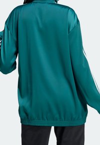 Henkilö, joka pitää yllään teal-väristä takkia, jossa on valkoiset raidat hihoissa, ja mustia housuja, kuvattu takaa.