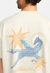 Crème-kleurig t-shirt met een grafiek van een blauwe gevleugelde paard, omringd door oranje en beige abstracte vormen op de achterkant.