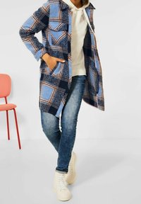 Abrigo a cuadros azul con patrones en navy y azul claro, que presenta un diseño de botones, dos bolsillos frontales y una textura suave, combinado con vaqueros y zapatillas blancas.