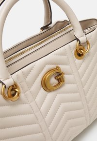 Sac à main en cuir matelassé crème avec fermeture éclair dorée, poignées doubles et émail "G" doré de la marque Guess à l'avant.
