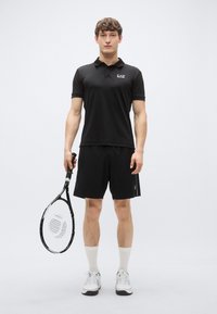 Svart polo med krage og logo, kombinert med svarte shorts. Modellen holder en tennisracket og har på seg hvite sokker og sko.