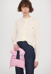 Krämfärgad knäpp cardigan med rund hals, i kombination med höga mörka jeans. Håller en ljusrosa tygväska med rynkad textur.