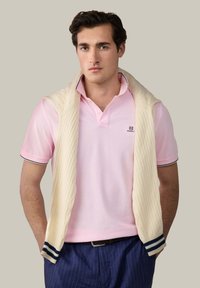 Mladý muž, ktorý má na sebe svetloružové polo tričko, námornícke nohavice s pruhmi a krémový sveter prehodený cez ramená.