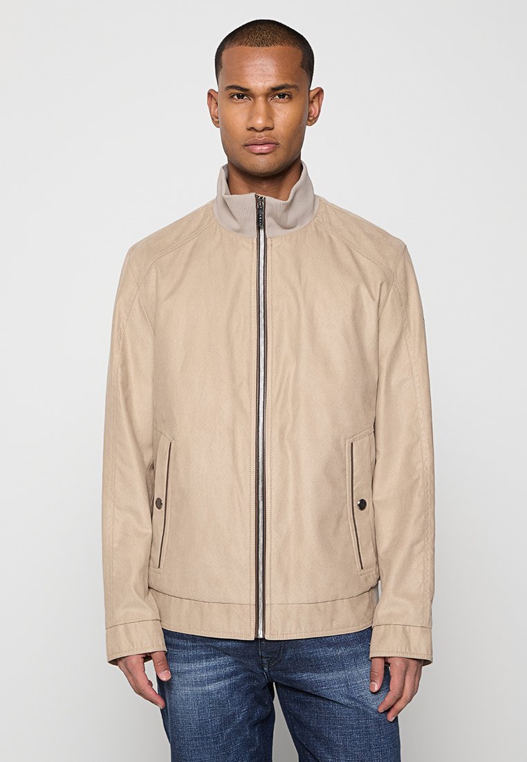 Veste beige en tissu léger, dotée d'un col haut et d'une fermeture éclair sur le devant. Deux poches latérales avec boutons et des lignes épurées.