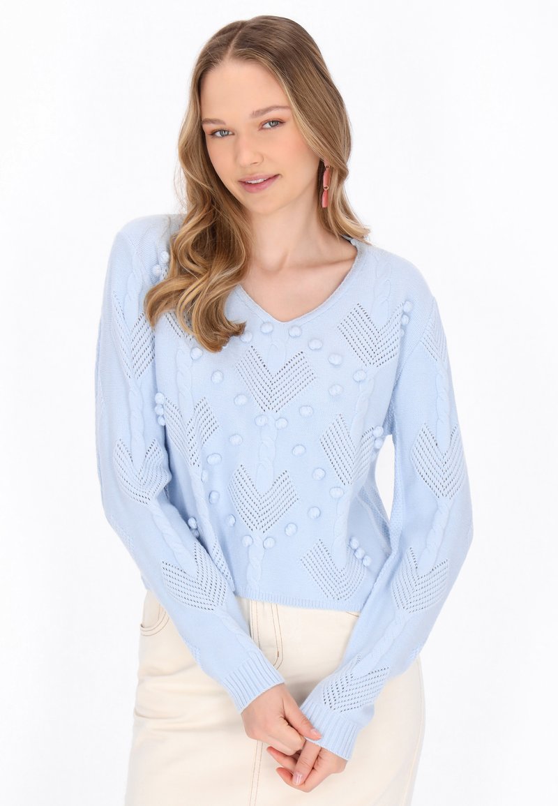 Maglione lavorato a maglia azzurro chiaro con scollo a V, caratterizzato da motivi testurizzati, dettagli a pompon e maniche lunghe. Abbinato a una gonna chiara.